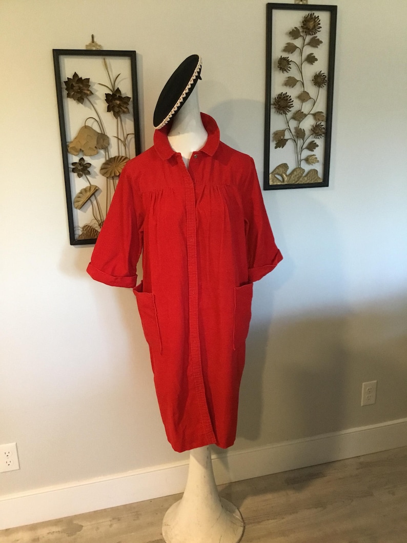 K&ouml;nnte beinhalten: Ein leuchtend rotes, kn&ouml;pfbares Kleid mit kurzen &Auml;rmeln und zwei Vordertaschen. Das Kleid wird auf einer wei&szlig;en Schaufensterpuppe pr&auml;sentiert. Ein schwarzer Hut mit wei&szlig;er Verzierung sitzt auf dem Kopf der Schaufensterpuppe. Wandkunst im Hintergrund.
