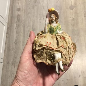 May include: A vintage porcelain figurine of a woman wearing a green and yellow dress and a hat. The figurine has a ruffled skirt made of fabric with a floral pattern. The figurine is held in a hand.
