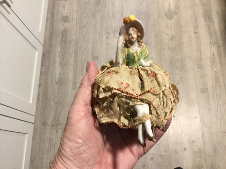 May include: A vintage porcelain figurine of a woman wearing a green and yellow dress and a hat. The figurine has a ruffled skirt made of fabric with a floral pattern. The figurine is held in a hand.