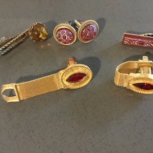 May include: A set of six gold-toned cufflinks and tie clips. The cufflinks feature a red stone and a pink patterned design. The tie clips feature a red stone and a pink patterned design. The tie clips are in the shape of a rectangle.