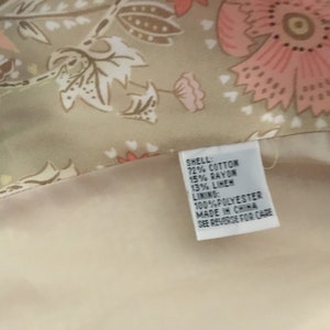 May include: A beige fabric with a floral print in shades of pink and brown. The fabric has a tag attached that reads "SHELL: 72% COTTON 15% RAYON 13% LINEN LINING: 100% POLYESTER MADE IN CHINA SEE REVERSE FOR CARE".