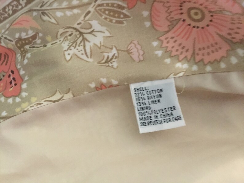 May include: A beige fabric with a floral print in shades of pink and brown. The fabric has a tag attached that reads "SHELL: 72% COTTON 15% RAYON 13% LINEN LINING: 100% POLYESTER MADE IN CHINA SEE REVERSE FOR CARE".
