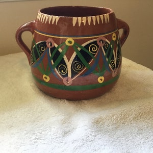 May include: A brown terracotta pot with two handles and a geometric design painted in black, green, blue, yellow, and pink.