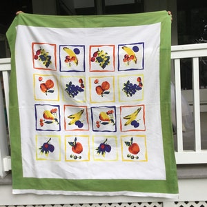 May include: A white tablecloth with a green border features a repeating pattern of colorful fruit illustrations in squares. The fruit includes pears, apples, grapes, and bananas.