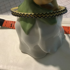 May include: A white ceramic figurine with a green and yellow fabric collar. The figurine is sitting on a white surface with a measuring tape.