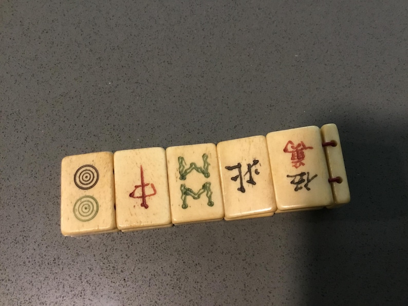 May include: A bracelet composed of five rectangular off-white tiles connected by thin brown cords. Each tile features distinct engraved markings, including concentric circles, a red vertical character, green symbols, and black Chinese characters.