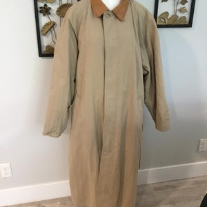 May include: A long, beige trench coat featuring a contrasting tan corduroy collar, long sleeves, and a concealed button-front placket. The coat has a straight, structured silhouette and reaches below the knees, providing full coverage.