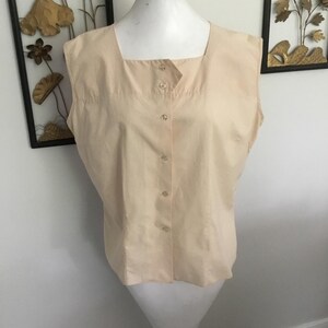 May include: A sleeveless, light peach-colored blouse with a square neckline and a button-down front. The blouse is displayed on a white mannequin. The background includes decorative wall art with floral designs.