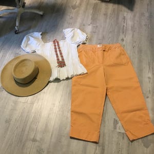 May include: A white short-sleeved blouse with a square neckline and gathered sleeves, a pair of orange cropped trousers with a button and zipper closure, a tan wide-brimmed straw hat, and a long beaded necklace with red and blue accents.