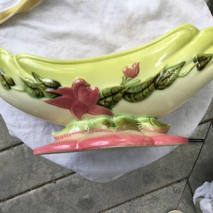 May include: A ceramic centerpiece bowl in a banana shape, with a pale yellow exterior and a pink base. The bowl is decorated with a red flower and green leaves. The bowl is approximately 14 inches long.