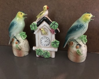 May include: A set of three vintage ceramic bird figurines. Two blue birds with yellow accents are perched on leafy bases. The third figurine is a white birdhouse with a clock face and a yellow bird on top.