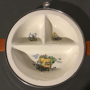 May include: A white ceramic divided plate with a silver rim and orange handles. The plate has three sections, each with a different cartoon image. One section has a white sheep, another has a person in a yellow hat, and the third has a pumpkin.