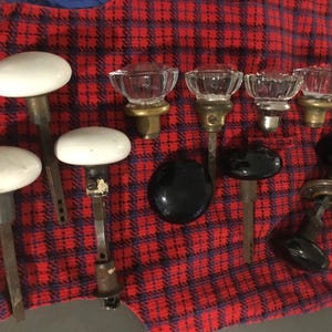May include: A collection of vintage door knobs in various styles. Includes white ceramic oval knobs, clear glass knobs, and black round knobs. The knobs are arranged on a red and black plaid fabric.