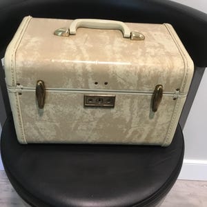 May include: A vintage cream-colored vanity case with a marbled pattern. The case features a white handle, gold-toned hardware, and a secure lock. The rectangular case is approximately 12 inches wide, 8 inches tall, and 7 inches deep.