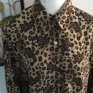 May include: A button-down shirt with a tan leopard print background and dark brown paisley designs. The shirt has a collared neckline and long sleeves. The buttons are white.