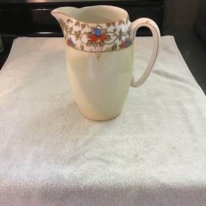 May include: A white ceramic pitcher with a brown, blue, and red floral design around the top. The pitcher has a wide mouth and a handle.