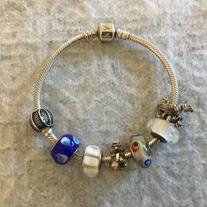 Peut inclure: Un bracelet à breloques en chaîne serpent argentée avec un fermoir central gravé du texte PANDORA. Le bracelet est orné de plusieurs breloques, dont une perle en verre bleu, une perle rayée blanche, un bonhomme de neige en métal, une perle multicolore et une grenouille.