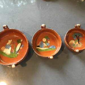 May include: Three terracotta bowls with painted designs. The largest bowl features a person in a blue shirt and a straw hat, the middle bowl has a house and a tree, and the smallest bowl has a tree with blue leaves.