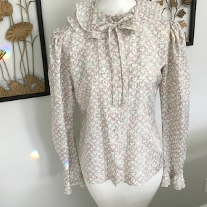 May include: A white blouse with a pink floral pattern and a ruffled collar. The blouse has long sleeves and a button-up front.