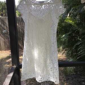 May include: A white lace dress with a floral pattern hangs on a hanger. The dress has short sleeves and a knee-length hemline. The intricate lace design covers the entire dress, creating a delicate and elegant look. The dress is hanging outside in natural light.