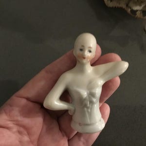 May include: A small, porcelain doll torso with a pale complexion, painted facial features, and a detailed bodice. The doll's arms are outstretched, and it has a small, rounded base. The doll is held in a hand.