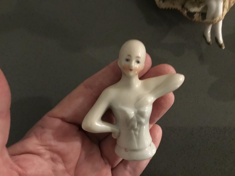 May include: A small, porcelain doll torso with a pale complexion, painted facial features, and a detailed bodice. The doll's arms are outstretched, and it has a small, rounded base. The doll is held in a hand.
