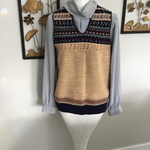 May include: A vintage sweater vest in tan, navy, and red. The vest has a V-neck and a patterned design across the shoulders. It is layered over a light blue blouse. The vest is displayed on a mannequin.