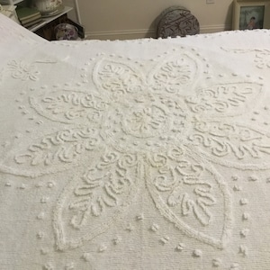 May include: A white cotton bedspread with a large, raised, floral design. The design is a stylized flower with a central circle and five petals.
