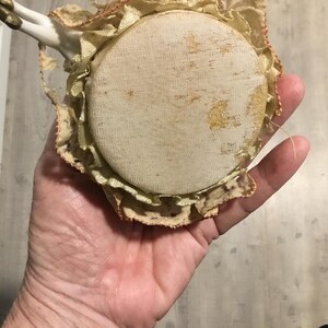 May include: A vintage pincushion with a round, cream-colored fabric top, showing signs of age. The pincushion is surrounded by a ruffled, lace-trimmed edge in shades of cream and gold. Small porcelain doll arms are attached.