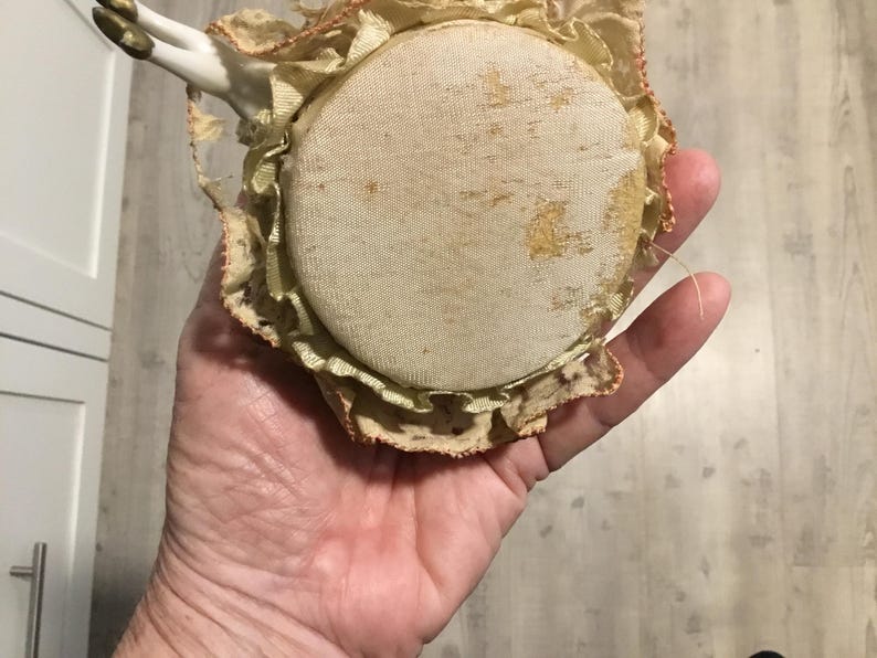 May include: A vintage pincushion with a round, cream-colored fabric top, showing signs of age. The pincushion is surrounded by a ruffled, lace-trimmed edge in shades of cream and gold. Small porcelain doll arms are attached.
