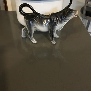 May include: A ceramic cow creamer with a black and white glaze, standing on a gray surface. The cow creamer has a curved handle and a spout for pouring. A white enamelware pot with a black rim and handle is in the background.