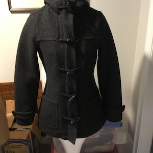 May include: A charcoal gray duffel coat with a hood and toggle closures. The coat has two front pockets and a blue lining visible at the hood. The coat is displayed on a white mannequin.