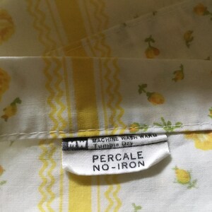Peut inclure: Un tissu blanc orn&eacute; d'un motif floral jaune avec des rayures verticales ondul&eacute;es jaunes. Une &eacute;tiquette d'entretien blanche est fix&eacute;e, affichant le texte MW, MACHINE WASH WARM, Tumble Dry, PERCALE et NO-IRON en noir sur fond blanc.