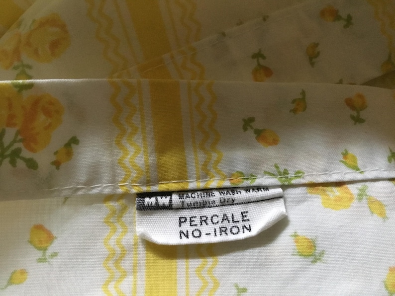 Peut inclure: Un tissu blanc orn&eacute; d'un motif floral jaune avec des rayures verticales ondul&eacute;es jaunes. Une &eacute;tiquette d'entretien blanche est fix&eacute;e, affichant le texte MW, MACHINE WASH WARM, Tumble Dry, PERCALE et NO-IRON en noir sur fond blanc.
