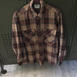 May include: A plaid button-down shirt in shades of tan, red, and navy blue. The shirt has two chest pockets and a collared neckline. The fabric appears to be a medium-weight material, suitable for cooler weather. The shirt is hanging against a dark green background.