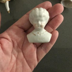 May include: A small, white porcelain doll head with a detailed face and curly hair. The doll head is a bust, with a collar and a hint of gold paint. The doll head is a vintage item.