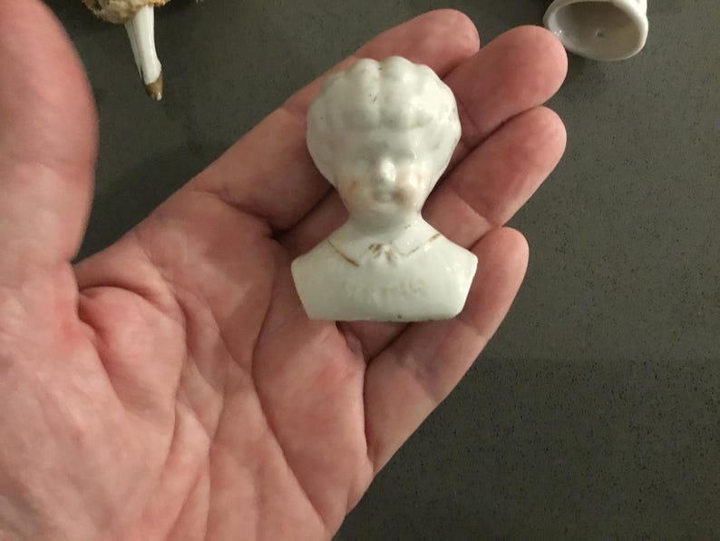 May include: A small, white porcelain doll head with a detailed face and curly hair. The doll head is a bust, with a collar and a hint of gold paint. The doll head is a vintage item.
