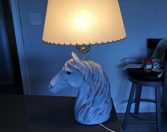 May include: A vintage table lamp featuring a white horse head base and a cream-colored lampshade. The lampshade has a scalloped edge and is lit from within. The lamp is on a dark surface.