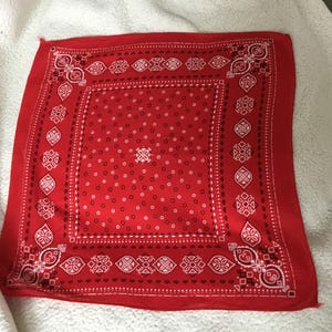 May include: Red bandana with a white paisley pattern and dotted accents. The square cloth features a central design and decorative borders. The bandana is displayed on a white, textured surface.