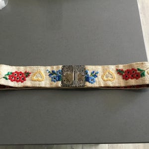 May include: Embroidered belt with a cream-colored woven base. The belt features red and blue floral designs, yellow heart shapes, and a silver-colored metal buckle. The belt is approximately 2 inches wide.