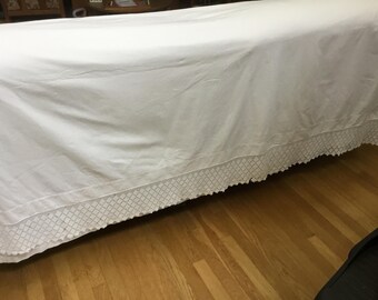 May include: A white bedspread with a decorative patterned border. The bedspread is draped over a bed, with the patterned border visible along the bottom edge. The bed is on a hardwood floor.