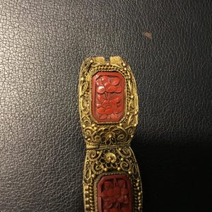 May include: A gold-toned metal filigree bracelet link featuring two rectangular, deep red carved floral inserts. The intricate wirework surrounds each red panel, which displays a detailed relief of blooming flowers and leaves in a symmetrical pattern.