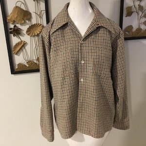 May include: A long-sleeved button-down shirt with a brown, black, and orange checkered pattern. The shirt has a wide collar, a single chest pocket, and is displayed on a white mannequin. The shirt is a vintage style.