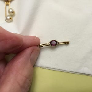 May include: A gold-toned bar pin with a central oval-shaped, deep pink gemstone. The pin is held against a white cloth, showcasing its delicate design. A second pin with pearls is partially visible in the background.