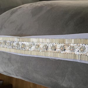 May include: A decorative belt featuring a central row of circular embellishments with clear and amber-colored stones, surrounded by silver-colored beads and sequins. The belt is set on a light gray fabric.