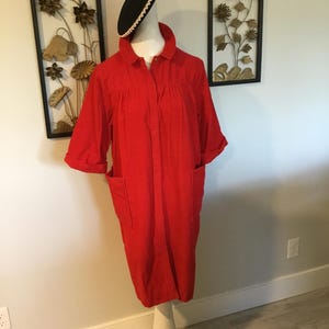 K&ouml;nnte beinhalten: Ein leuchtend rotes Kleid mit kurzen &Auml;rmeln und zwei Vordertaschen. Das Kleid hat Kn&ouml;pfe und einen Kragen. Ein schwarzer Hut mit wei&szlig;er Bord&uuml;re sitzt auf dem Kopf der Schaufensterpuppe. Das Kleid wird auf einem wei&szlig;en Mannequin-St&auml;nder pr&auml;sentiert.