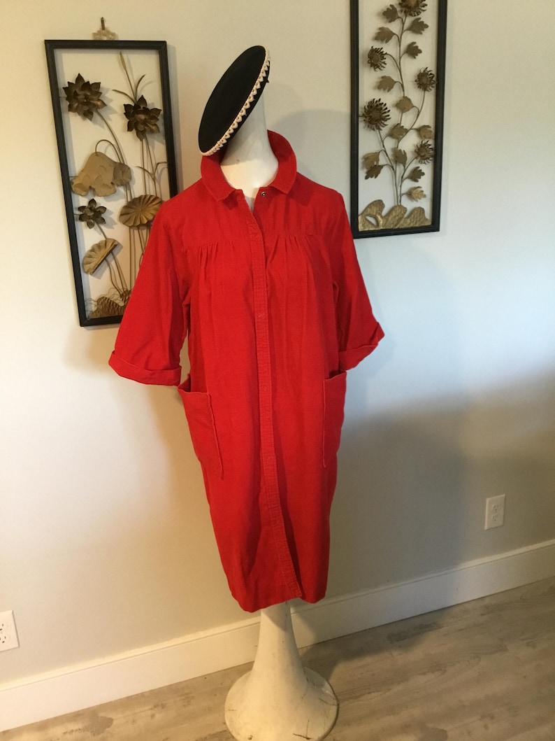 K&ouml;nnte beinhalten: Ein leuchtend rotes Kleid mit kurzen &Auml;rmeln und zwei Vordertaschen. Das Kleid hat Kn&ouml;pfe und einen Kragen. Ein schwarzer Hut mit wei&szlig;er Bord&uuml;re sitzt auf dem Kopf der Schaufensterpuppe. Das Kleid wird auf einem wei&szlig;en Mannequin-St&auml;nder pr&auml;sentiert.