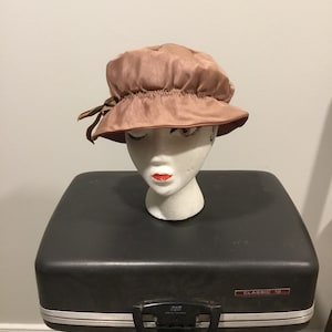 May include: A vintage pink satin hat with a gathered crown and a brown ribbon bow. The hat is displayed on a white mannequin head with red lipstick, placed on a black suitcase. The suitcase is labeled "CLASSIC 12".