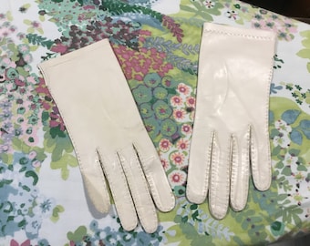 May include: A pair of off-white leather gloves with a stitched design. The gloves are laid out on a floral fabric background.