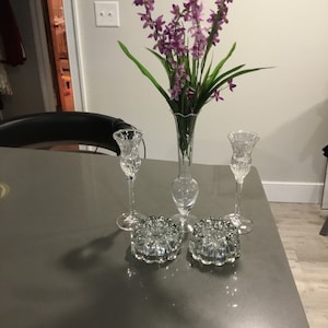 May include: Two clear glass candle holders with a decorative base and a bouquet of purple flowers in a clear glass vase.
